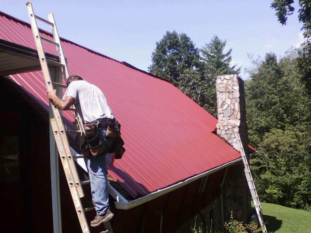 Roofer on a ladder beside a red metal roof and stone chimney — Soffit & Fascia Repair by Roofing Brothers in Bethesda